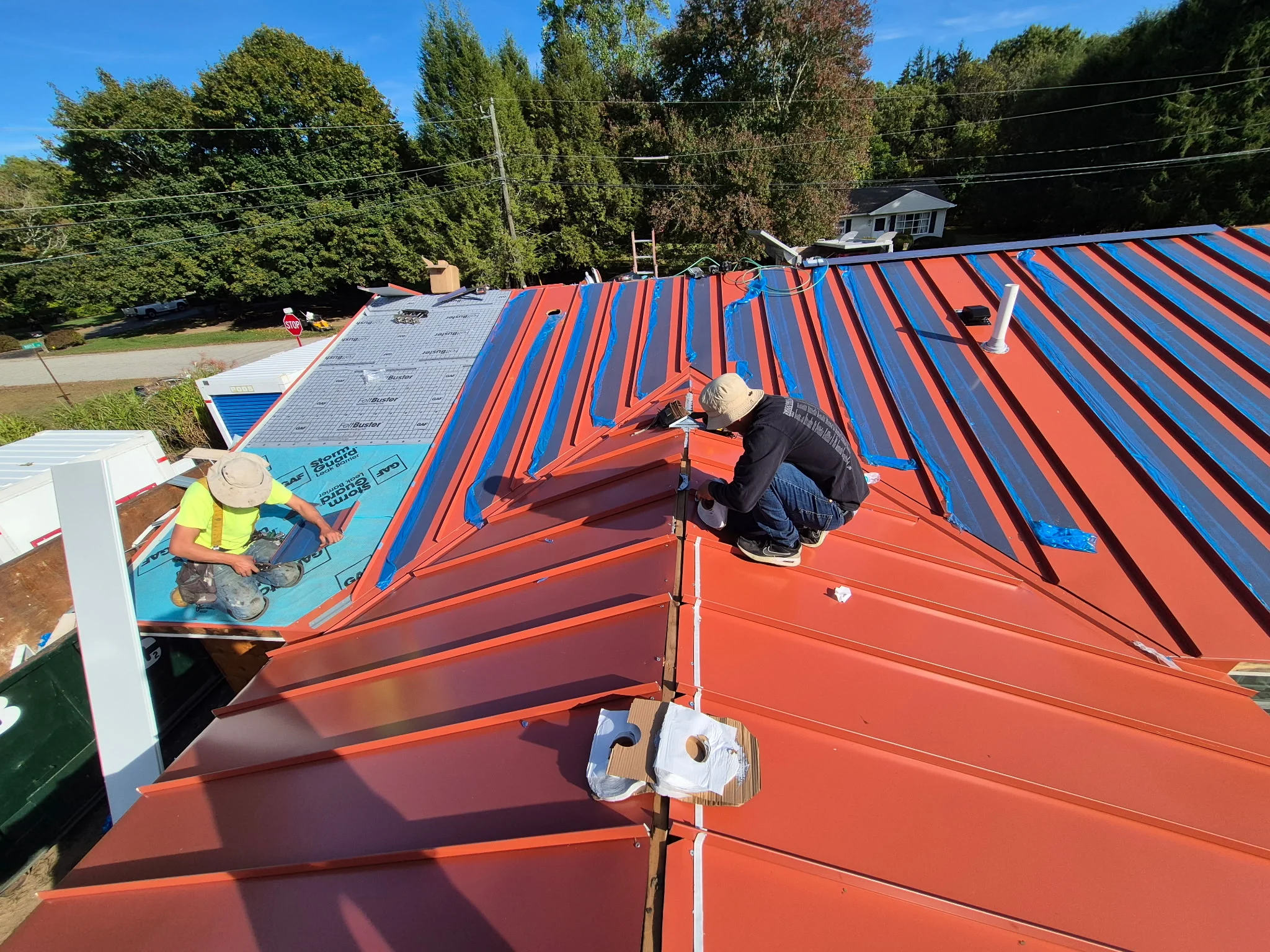 Metal standing seam roof installation with orange and blue panels, workers installing fasteners