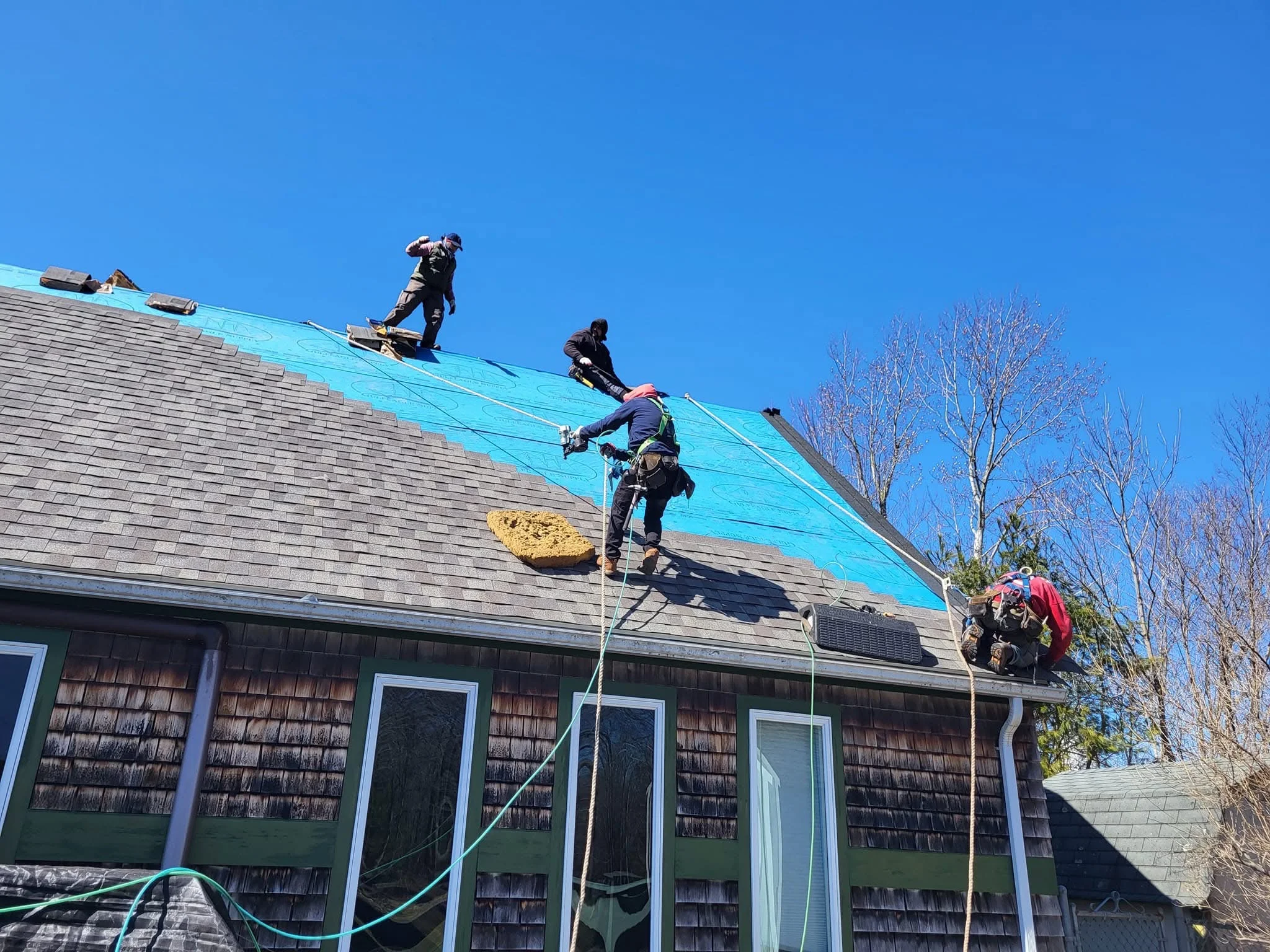 Roofers installing gray and blue architectural shingles on residential roof with safety equipment